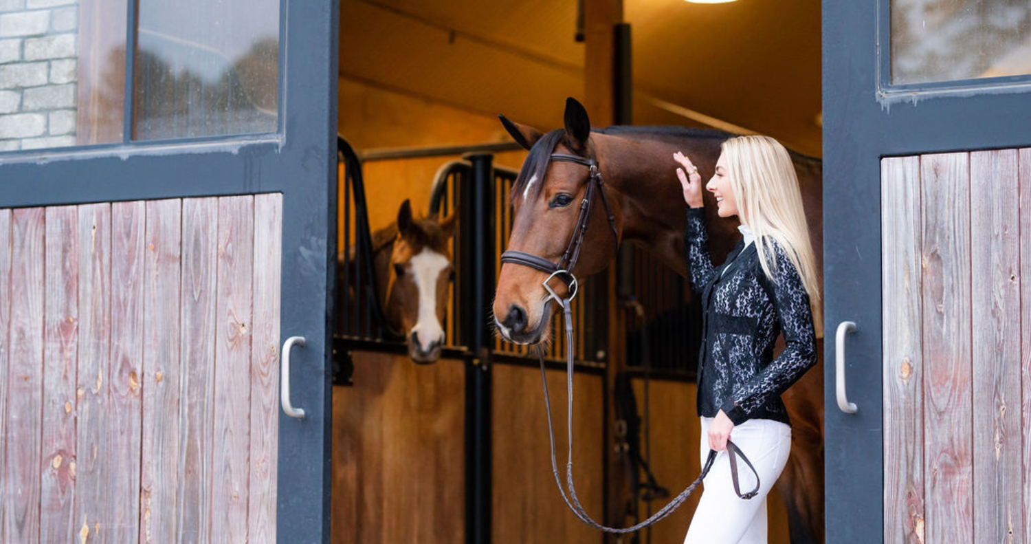 blonde woman wearing lace show coat stands with bay horse in between barn doors. equestrian clothing clearance, horse riding clothes, horse clothing